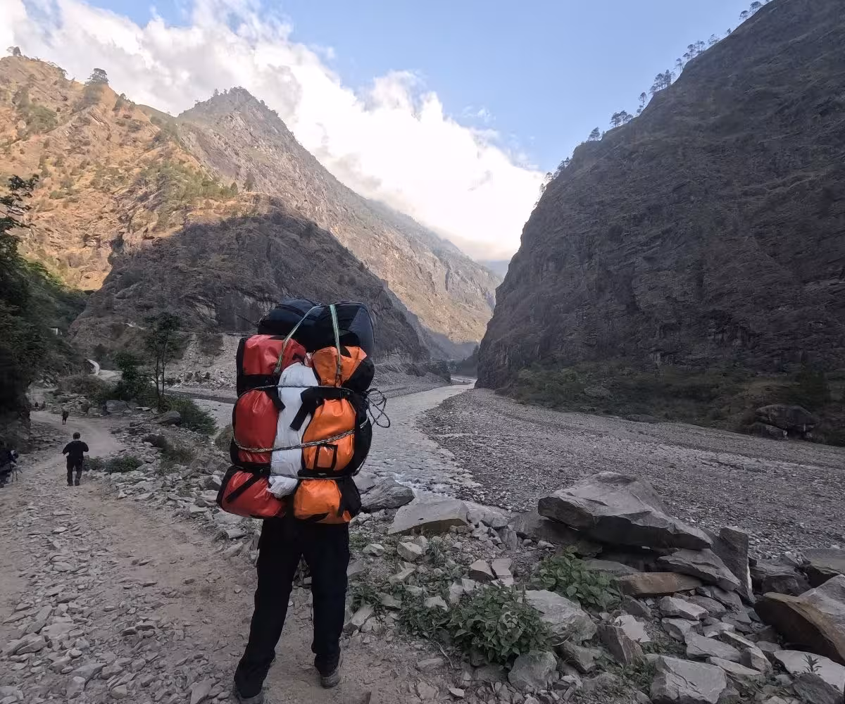 trek porter walking beside a river with large backpacks tied together
